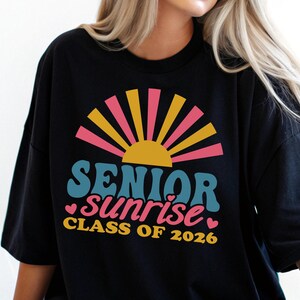 Puede incluir: Camiseta negra con un dise&ntilde;o retro que presenta las palabras "SENIOR sunrise" en verde azulado y rosa, con un gr&aacute;fico de sol amarillo y rosa. Tambi&eacute;n se incluye el texto "CLASS OF 2026".
