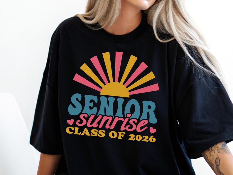 Puede incluir: Camiseta negra con un dise&ntilde;o retro que presenta las palabras "SENIOR sunrise" en verde azulado y rosa, con un gr&aacute;fico de sol amarillo y rosa. Tambi&eacute;n se incluye el texto "CLASS OF 2026".