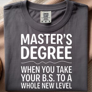 Può includere: Una maglietta grigia con le parole "MASTER'S DEGREE" in grandi lettere bianche. Sotto, il testo recita "WHEN YOU TAKE YOUR B.S. TO A WHOLE NEW LEVEL". La maglietta ha uno scollo tondo e un'etichetta di Comfort Colors.