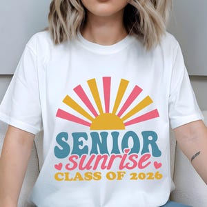 Puede incluir: Camiseta blanca con un dise&ntilde;o gr&aacute;fico colorido. El dise&ntilde;o incluye un sol estilizado con rayos rosas y amarillos, y las palabras "SENIOR sunrise CLASS OF 2026" en verde azulado, rosa y amarillo. Tambi&eacute;n se incluyen peque&ntilde;os corazones rosas.