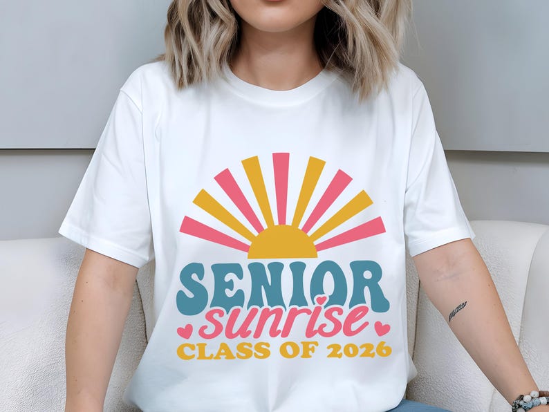 Puede incluir: Camiseta blanca con un dise&ntilde;o gr&aacute;fico colorido. El dise&ntilde;o incluye un sol estilizado con rayos rosas y amarillos, y las palabras "SENIOR sunrise CLASS OF 2026" en verde azulado, rosa y amarillo. Tambi&eacute;n se incluyen peque&ntilde;os corazones rosas.