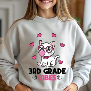 Puede incluir: Sudadera gris claro con un gato de dibujos animados con gafas rosas y una burbuja de chicle. Corazones rosas rodean al gato, con el texto "3RD GRADE VIBES" en negro y rosa debajo. Un estilo casual y cómodo.