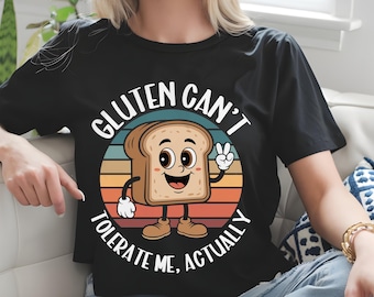 May include: Black t-shirt featuring a cartoon bread slice character within a colourful circular design. The text "GLUTEN CAN'T TOLERATE ME, ACTUALLY" is printed around the bread. The bread slice is smiling and giving a peace sign.