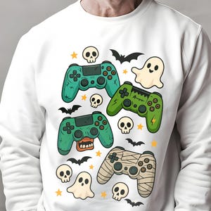 May include: White sweatshirt featuring a Halloween-themed design. The graphic includes green and tan game controllers, skulls, bats, ghosts, and stars. The controllers have a spooky design, with one wrapped in bandages.