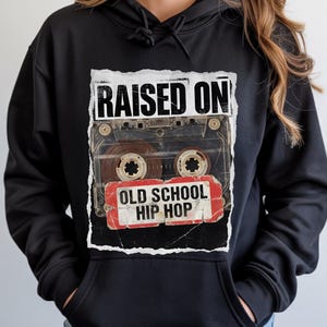 May include: Black hoodie featuring a graphic of a cassette tape with the words "RAISED ON" above and "OLD SCHOOL HIP HOP" below. The design has a vintage, distressed aesthetic, with a torn paper effect.