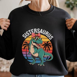 May include: Black sweatshirt featuring a graphic of a green dinosaur wearing sunglasses and a red bow, with the text "SISTERSAURUS" above. The dinosaur is set against a sunset background with palm trees.