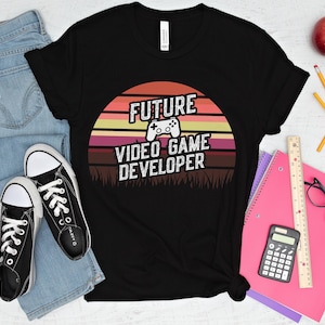 Peut inclure: T-shirt noir avec l'inscription "FUTURE VIDEO GAME DEVELOPER" et un motif de manette de jeu, sur fond de coucher de soleil. Le t-shirt est associé à un jean bleu et des baskets noires. On y trouve aussi des fournitures scolaires.