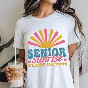 Puede incluir: Camiseta blanca con un dise&ntilde;o gr&aacute;fico colorido. El dise&ntilde;o presenta un sol naciente con rayos rosas y amarillos, y las palabras "SENIOR sunrise CLASS OF 2026" en verde azulado y rosa. La persona sostiene un vaso de caf&eacute; helado.