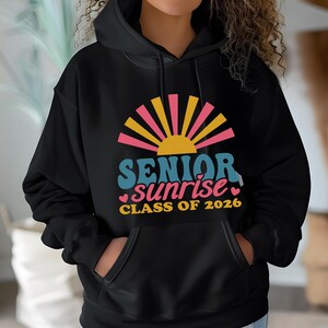 Puede incluir: Sudadera con capucha negra con un gr&aacute;fico colorido. El dise&ntilde;o presenta un sol naciente con las palabras "SENIOR sunrise CLASS OF 2026" en turquesa, rosa y amarillo. La sudadera tiene un bolsillo delantero y una capucha con cord&oacute;n.