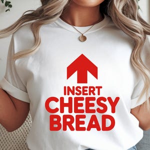 May include: White t-shirt with a red arrow pointing up and the words "INSERT CHEESY BREAD" in red, bold letters. The shirt has short sleeves and a crew neck.