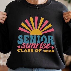 Puede incluir: Sudadera negra con un dise&ntilde;o gr&aacute;fico colorido. El dise&ntilde;o presenta un sol estilizado con rayos rosas, amarillos y naranjas, y las palabras "SENIOR sunrise CLASS OF 2026" en azul, rosa y amarillo.