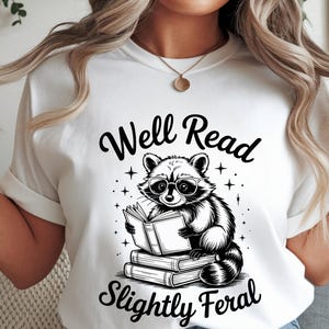 May include: White t-shirt featuring a black and white illustration of a raccoon wearing glasses and reading a book. The text "Well Read Slightly Feral" is above and below the raccoon image. The shirt has short sleeves.