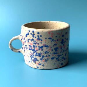 Könnte beinhalten: Ein Set aus drei Keramikbechern mit gesprenkelter, matter Oberfläche. Jeder Becher hat eine andere Farbe: hellblau, cremefarben und grau. Die Becher haben ein schlichtes, modernes Design und einen bequemen Griff.