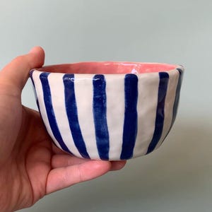 Könnte beinhalten: Eine kleine, runde Keramikschale mit einem handbemalten Design aus vertikalen marineblauen Streifen auf weißem Grund. Das Innere der Schale ist mit einer einfarbigen, leuchtend rosa Glasur versehen, und die Außenfläche hat eine leicht unebene Textur.