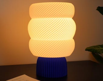 Könnte beinhalten: Eine Tischleuchte mit einem leuchtenden, weißen, gerippten Glasschirm in einem dreistufigen, abgerundeten Design mit Spiralstruktur. Der Lampenfuß ist ein massives, tiefblaues, zylindrisches Element mit vertikalen Rillen als stabiler Stand.