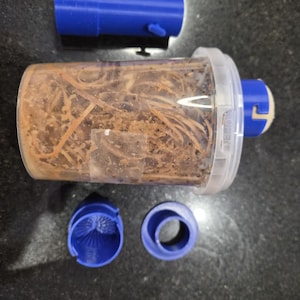 May include: A clear plastic fruit fly trap container filled with brown organic material, featuring a white lid with the word OPEN printed on it. The set includes three separate blue plastic components: a cylindrical tube and two circular caps.