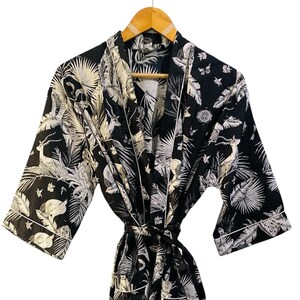 May include: A black and white patterned robe with a tie belt, hanging on a wooden hanger. The robe features a botanical and animal print design with white trim along the edges.