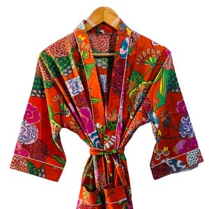 Op de afbeelding: Een feloranje badjas met een kleurrijk bloemenpatroon. De badjas heeft een strik in de taille, wijde mouwen en witte biezen. Het ontwerp bevat roze, groene, blauwe en gele bloemelementen, waardoor een levendig kledingstuk ontstaat.