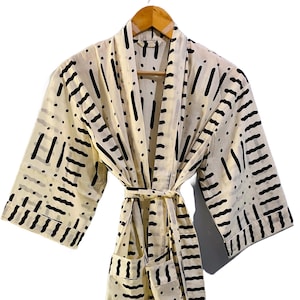 May include: Cream-colored robe with a black geometric pattern. The robe has a matching belt, pockets, and wide sleeves. It is hanging on a wooden hanger against a white background. The robe is made of a lightweight fabric.
