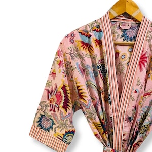 Op de afbeelding: Een roze badjas met bloemenprint en bijpassende ceintuur, hangend aan een houten hanger. De badjas heeft een kleurrijk patroon van bloemen en bladeren in blauw, geel en rood. De mouwen en de bies hebben een gestreept ontwerp.