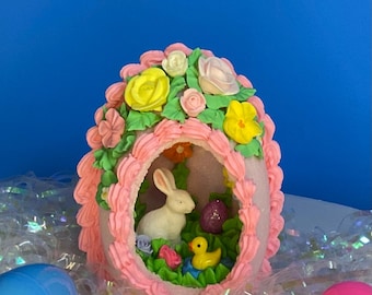 May include: A pink Easter egg-shaped decoration with a white rabbit, yellow duck, and purple egg inside. The egg is decorated with pink frosting and colorful flowers. Several colorful Easter eggs surround the decoration.