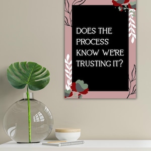 Op de afbeelding: Een ingelijste kunstdruk met de tekst "DOES THE PROCESS KNOW WE'RE TRUSTING IT?" op een zwarte achtergrond, omlijst in roze met bloemen- en bladaccenten. Een heldere glazen vaas met een groen blad en een kleine witte kom staan op een wit oppervlak.