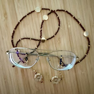 May include: A pair of gold-framed spectacles with clear lenses, accompanied by a beaded eyeglass chain. The chain is made of small dark red beads, gold-coloured accents, and decorative flower charms. The spectacles are placed on a wooden surface.