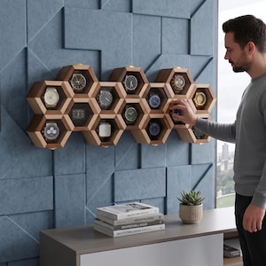 May include: A wooden watch display case with multiple hexagonal compartments, each holding a wristwatch. The case is mounted on a blue wall. A person is reaching for a watch. Books and a small plant are on a nearby table.