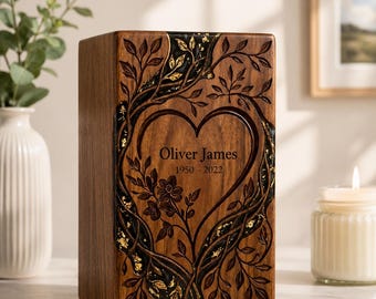 May include: A rectangular wooden cremation urn featuring a carved heart shape surrounded by intricate tree branches and leaves. The design includes black resin accents with gold leaf flakes. The text reads Oliver James 1950-2022.
