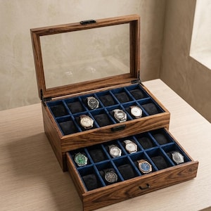 Puede incluir: Una caja de madera para exhibición de relojes con tapa de cristal transparente, abierta para revelar múltiples relojes. El interior presenta un forro de terciopelo azul y compartimentos individuales para cada reloj. La caja está sobre una superficie de madera clara.
