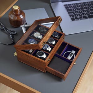 Puede incluir: Una caja de reloj de madera con tapa de cristal transparente, que muestra varios relojes de pulsera con varios diseños de correa y esfera. La caja tiene un cajón con una cartera y otros accesorios. El interior está forrado con terciopelo azul.