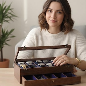 May include: A dark wooden watch display case with a glass lid, displaying multiple watches. The case has a dark brown finish and a blue velvet interior. The drawer is open, revealing extra storage. The case is on a wooden table.