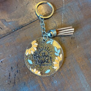 May include: A clear acrylic keychain with a gold ring and chain, featuring a round charm with the text "Be the Light" and floral designs in yellow and blue. A tan tassel hangs from the charm.