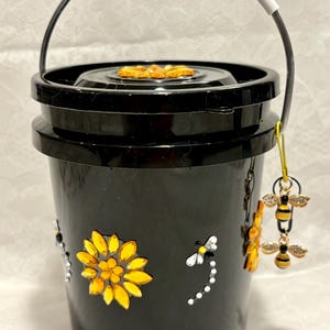 May include: A black plastic bucket with a white handle, decorated with yellow and clear crystal sunflower and dragonfly embellishments. A gold-toned charm featuring two small bee figures hangs from the handle on a yellow cord.