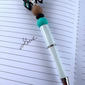 May include: A decorative pen with a white barrel and silver accents. The pen features a turquoise, black, and brown beaded design, topped with a detailed, colorful bull skull charm. The pen rests on a lined notebook page with the word "Love" written in cursive.