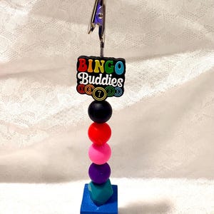 May include: A metal clip holder featuring a black rectangular charm with the text Bingo Buddies and colorful bingo ball icons. The stem consists of stacked round beads in black, red, pink, and purple, topped with a teal geometric bead.