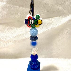 May include: A metal alligator clip attached to a vertical stack of beads in various shades of blue, including textured, frosted, and clear finishes. At the top, a white charm features the word BINGO with colorful numbered bingo balls.