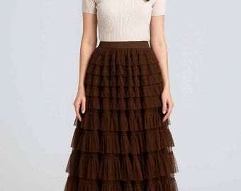 May include: A woman wearing a cream-colored short-sleeved top and a long, tiered brown tulle skirt. The skirt has multiple layers of ruffled fabric. She is also wearing nude-colored heels.