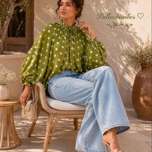 Puede incluir: Una blusa verde oliva con lunares blancos, con cuello alto con volantes, mangas largas voluminosas con puños fruncidos y un detalle de lazo frontal. La blusa se combina con pantalones vaqueros anchos de color azul claro y sandalias de tacón beige.