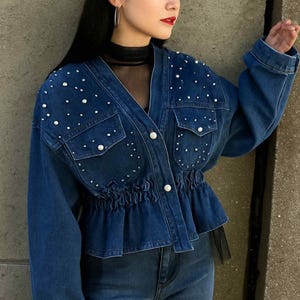 May include: A dark wash denim jacket with pearl embellishments and a cinched waist. The jacket features two front pockets and button closures. The model is also wearing matching denim jeans.