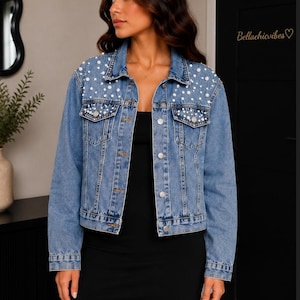 Puede incluir: Chaqueta vaquera azul claro adornada con perlas blancas. La chaqueta tiene un diseño clásico con cierre de botones, bolsillos en el pecho y cuello. El texto "Bellachicvibes" es visible en la puerta.