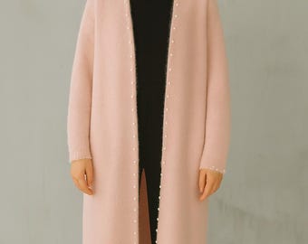 May include: A long, pale pink cardigan with a soft, fuzzy texture. The cardigan is adorned with small, white pearl-like beads along the edges. The open front design reveals a black top and pants underneath, creating a layered look.