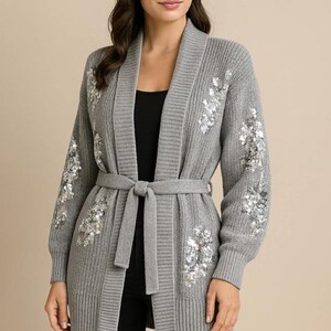 May include: A grey knitted cardigan with a shawl collar and a matching belt. The cardigan has floral sequin embellishments on the sleeves and front. The model is wearing a black top and black trousers.
