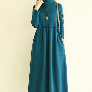 Könnte beinhalten: Ein teal-farbenes, langärmliges Kleid mit Rollkragen und fließendem Rock. Das Kleid hat eine betonte Taille, Taschen und eine lange Halskette. Braune Schuhe vervollständigen das Outfit. Die Länge beträgt ca. 130 cm.