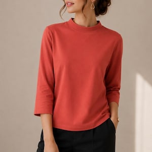 May include: A terracotta-coloured top with a mock neck and three-quarter length sleeves, paired with black trousers. The garment features a solid, smooth fabric construction with a clean, straight hemline and a simple, unadorned design throughout.