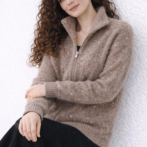May include: A light brown zip-up sweater with a high collar and subtle sequin embellishments. The sweater is paired with a black skirt. The model is sitting against a white wall.