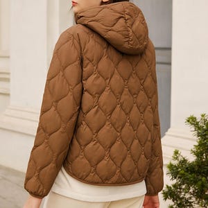 May include: A brown quilted jacket with a hood. The jacket features a diamond pattern and is worn over a white top and beige trousers. The model is standing outside near a building and some greenery.