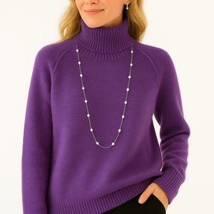 Pode incluir: Uma mulher vestindo um suéter roxo de gola alta e um colar de pérolas longo. O suéter tem mangas compridas e bainha canelada. O colar é uma delicada fileira de pérolas. A mulher também usa brincos pequenos.