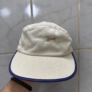 May include: A beige cap with a navy blue brim. The cap features the words "stussy Sport" embroidered in brown. The cap is made of a textured fabric.