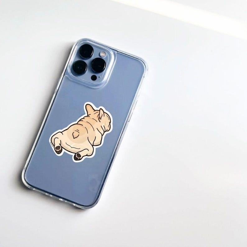 K&ouml;nnte beinhalten: Eine transparente Handyh&uuml;lle mit einem hellblauen Telefon im Inneren. Auf der R&uuml;ckseite des Telefons befindet sich ein Aufkleber eines hellbraunen French Bulldogs in Ruheposition. Das Telefon hat drei schwarze Kameralinsen.